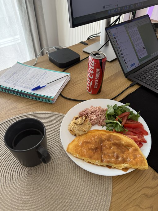 A home office desk setup with a dual monitor and laptop workspace, featuring a Coca-Cola Zero Sugar can, a black mug, a plate with an omelet, salad, tuna, and hummus, alongside a spiral notebook.