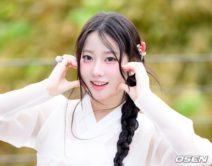 A smiling young woman with a long black braid wears a white traditional outfit, posing with her hands framing her face; the 'OSEN' watermark is visible in the bottom right corner.
