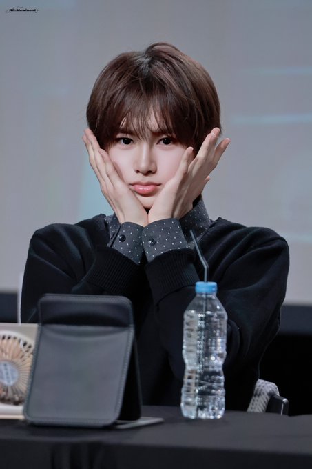 A young man with brown hair poses with his hands framing his face, wearing a black sweater over a polka-dot collared shirt, seated at a table with a water bottle and tablet in front of him—likely Anshin (周安信) of the K-pop group ALPHADRIVEONE (ALD1) during a fan event on September 21, 2026.