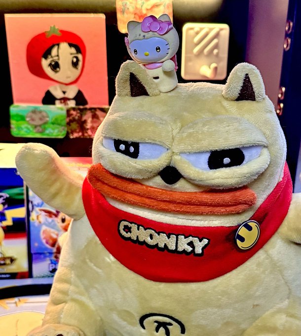 A plush toy named 'CHONKY' resembling a grumpy cat wears a red bandana with a Hello Kitty figurine on its head, displayed in front of a colorful gaming setup.