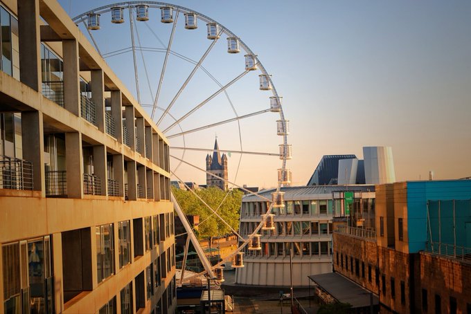 The image features an aerial view of a cityscape with tall buildings surrounding a large Ferris wheel. The Ferris wheel is situated between two high-rise buildings, creating a unique juxtaposition of architectural styles. The sky above appears hazy, adding a sense of depth and atmosphere to the scene. In the foreground, there are some trees visible, providing a touch of nature amidst the urban setting.