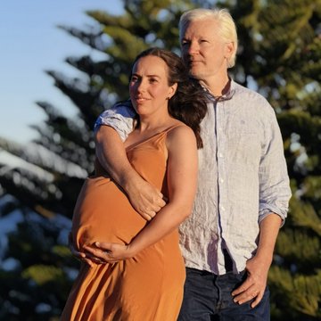 A man with long blonde hair embraces a pregnant woman in an orange dress as they stand together outdoors, with the woman's hands resting gently on her baby bump.

The image depicts Julian Assange with his wife, illustrating the post's message about his personal life journey—from being imprisoned to gaining freedom and now building a family with their expected third child.