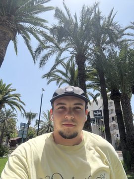 A young man with a goatee wearing a light yellow t-shirt and white backwards cap, taking a selfie in front of palm trees and a building on a sunny day.