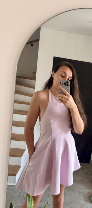 A woman takes a mirror selfie wearing a light pink halter dress, standing in a modern home with a curved staircase visible in the reflection.