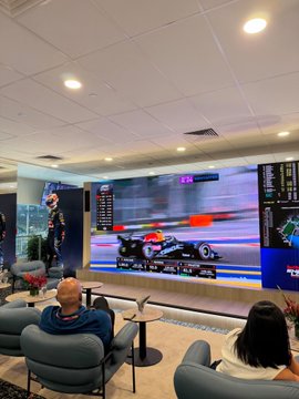 Two people sitting in a lounge area watching a Formula 1 race on a large curved screen, with race statistics displayed on the wall.