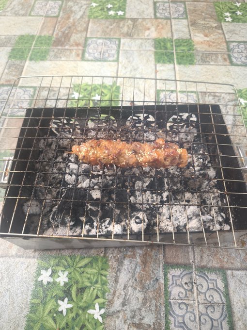 A skewer of meat grilling over hot coals on a small charcoal grill placed on a tiled floor.
