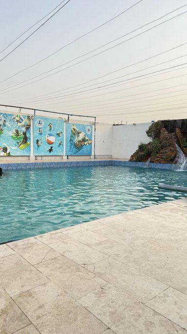 An outdoor swimming pool area with a light-colored tile deck, a shade sail overhead, a decorative wall mural, and a small rock waterfall feature.