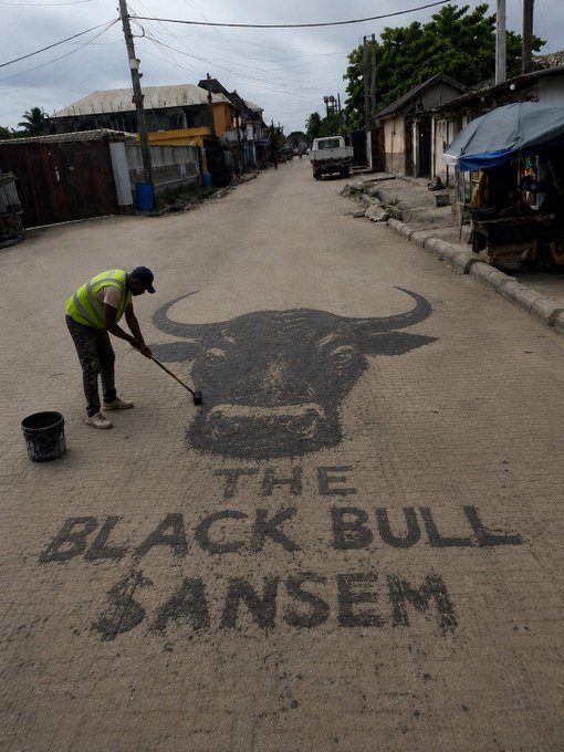A street artist in a yellow safety vest paints a large bull logo and the text "THE BLACK BULL BANSEM" on a quiet street, with a black bucket beside him.