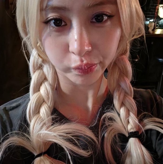 A close-up selfie of a person with blonde hair styled in twin braids, wearing dramatic winged eyeliner and a grey top.