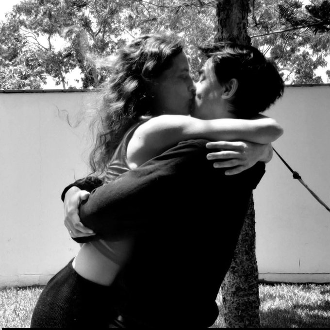A black-and-white photograph showing two women embracing and kissing outdoors next to a tree, with one wearing white fluffy ear accessories.