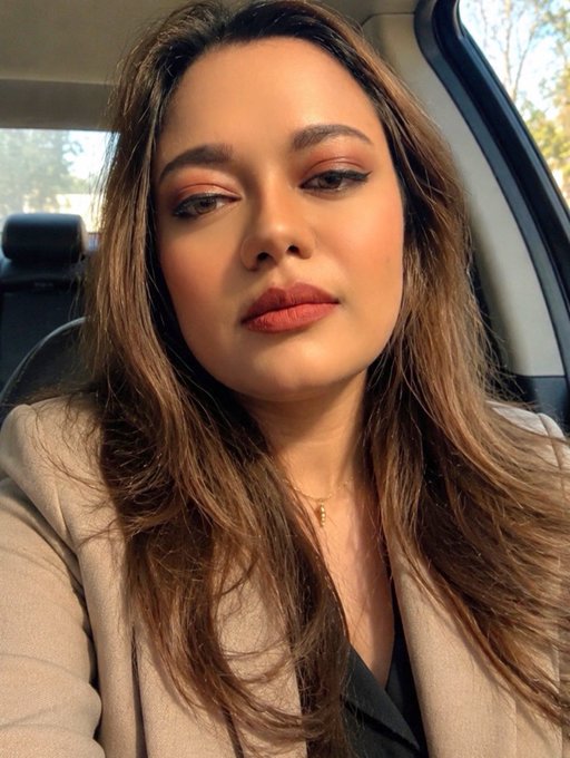 A woman with long brown hair, dramatic eye makeup, and matte lipstick takes a selfie while sitting in a car, wearing a beige blazer.