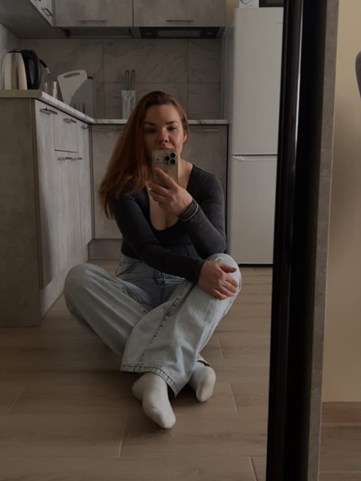 A mirror selfie of a young woman with long brown hair sitting cross-legged on a wooden kitchen floor, wearing a black long-sleeve top and light blue jeans, taking the photo with a smartphone.