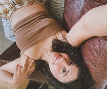 The image, presented sideways, shows a woman with dark hair wearing a brown top lying against a patterned cushion, playfully posing for the camera. The caption pairs her with the artwork she's modeling for, highlighting her as both the artist and the inspiration behind the piece available for mint.
