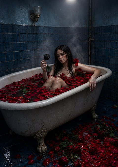 A woman with wet hair sits in a vintage clawfoot bathtub filled with water and red rose petals, holding a black rose in a dimly lit, blue-tiled bathroom with steam rising around her.