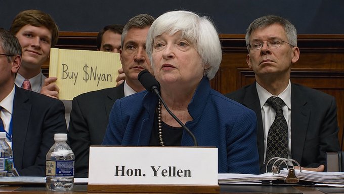 Treasury Secretary Janet Yellen, identified by a 'Hon. Yellen' nameplate, speaks at a congressional hearing while a young attendee behind her holds up a sign reading 'Buy $Nyan'.