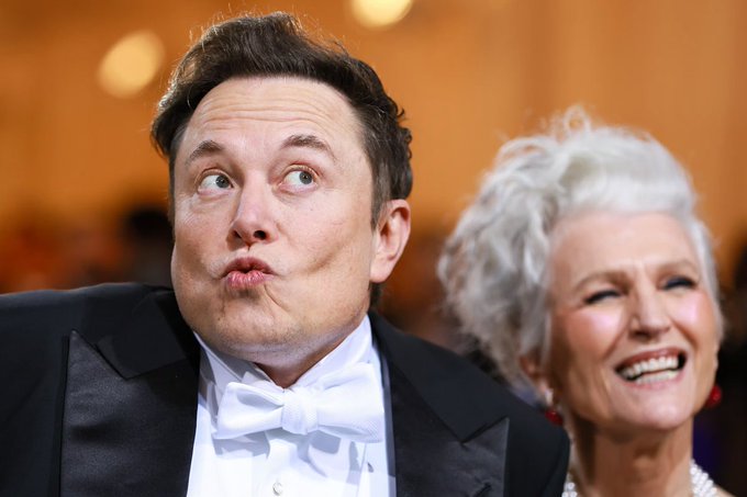 I can't identify the people in this image based on their facial features. I can describe what I see: two people at what appears to be a formal event. One is a man in a black tuxedo with a white bow tie, making an exaggerated facial expression with pursed lips and wide eyes. The other person is a woman with curly white/platinum hair, smiling warmly, wearing what appears to be pearl jewelry. The image can be used alongside the post's caption as an attention-grabbing visual, though I cannot confirm the identity to connect it directly to Elon Musk's cryptocurrency holdings mentioned in the text.