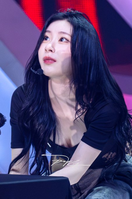 A young woman with long dark hair, wearing a black top and a headset microphone, looks upward while seated in what appears to be a brightly lit studio setting.