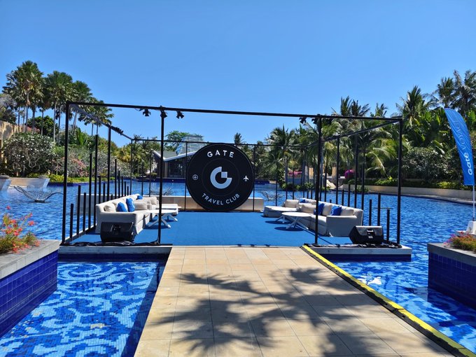 An outdoor event stage setup branded 'GATE 4 TRAVEL CLUB' positioned over a large blue swimming pool, featuring white lounge seating and surrounded by palm trees under a clear blue sky.