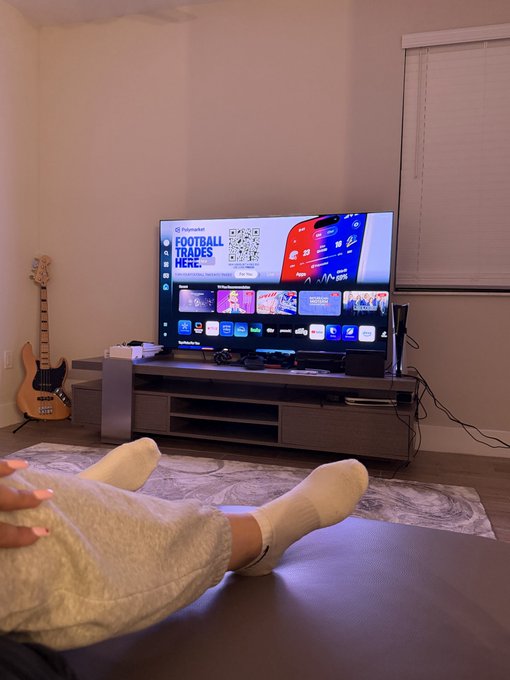 A first-person view shows a person's sock-clad feet resting in front of a large television displaying a Fanatics Football Trades menu, with a bass guitar leaning against the wall nearby.