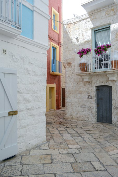 A narrow stone-paved alleyway is lined with whitewashed buildings, including a red-walled house and a stone building with blue shutters and a balcony adorned with pink flowers.