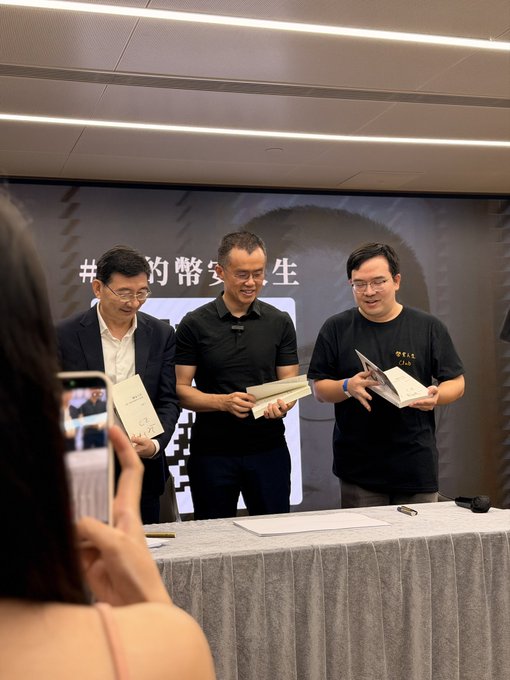 Three men in business casual attire stand at a table holding open books, with a partial hashtag containing '#的常态' visible on the backdrop behind them, while a person in the foreground photographs the scene with a phone.