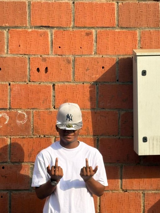 A young man wearing a white t-shirt and a New York Yankees cap (with the iconic "NY" logo) makes the middle finger gesture with both hands while standing against a red brick wall. The casual, defiant pose mirrors the post's playful and confident tone, reflecting his carefree attitude and good vibes from spending time with friends.