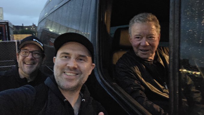 A selfie shows three men smiling together backstage at Riot Fest, with an older man visible through what appears to be a tour bus window, while the accompanying post identifies one of the men as William Shatner and describes the moment as a charity interaction captured for Elon Musk.