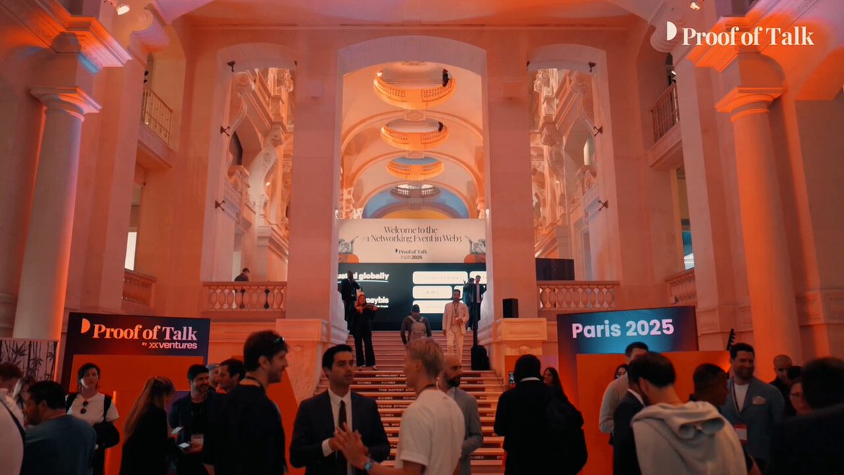 The image shows a large group of people gathered inside an ornate building, possibly attending a conference or event. The attendees are standing around a stage with a banner that reads "Proof of Talk" and "Bittensor". There are several people on the stage, some holding drinks in their hands. A few individuals can be seen walking around outside the main area of the building. The scene suggests an atmosphere of social interaction and networking among the attendees.