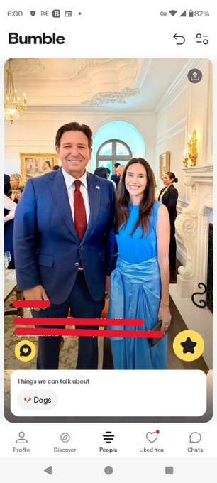 A Bumble dating app profile screenshot showing a man in a blue suit and red tie posing with a woman in a blue dress, with the bio redacted and the interests tag 'Dogs'.