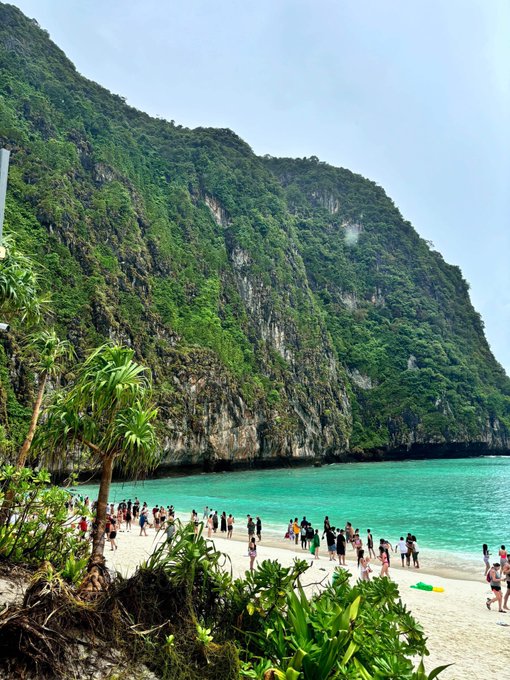 A tropical beach scene featuring turquoise waters, white sand crowded with tourists, and steep limestone cliffs covered in lush green vegetation.
