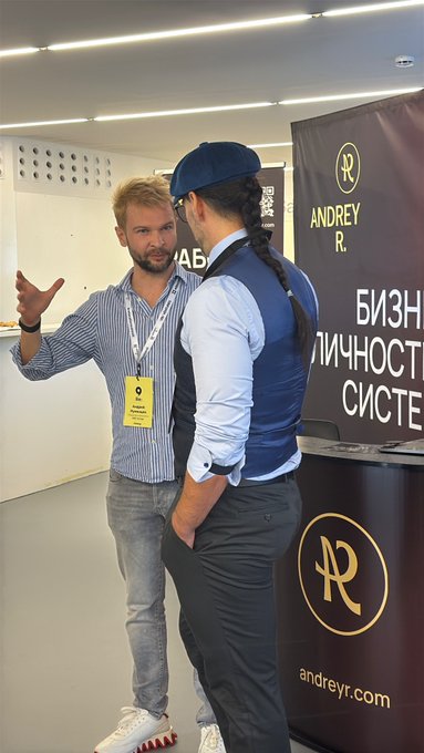 Two men talking at what appears to be a business event, with one wearing a striped shirt and lanyard, and the other in a blue vest and cap with a long braided ponytail, standing near an 'ANDREY R.' (andreyrok) banner with Russian text.