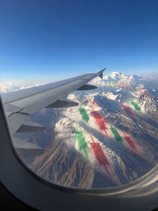 A view from an airplane window showing the wing and snow-capped mountains below, with colorful red and green banner-like overlays across the landscape.