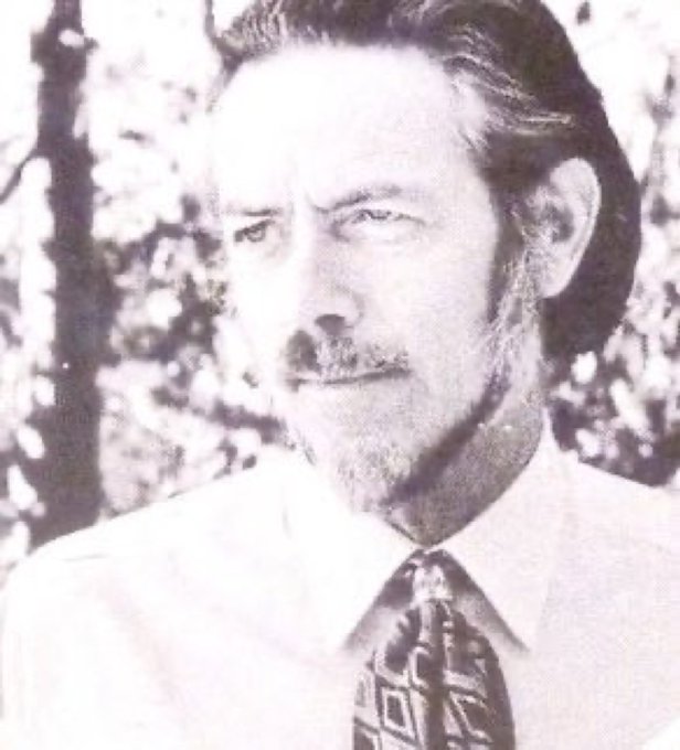 A vintage black-and-white portrait of a mustached man in a suit and patterned tie, set against a softly blurred outdoor background.