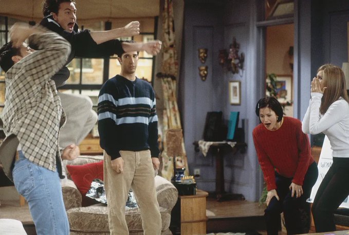 A scene from the TV show Friends showing the characters in Monica's apartment. Ross and Chandler appear to be jumping or fighting playfully in the background, while Joey stands looking stunned in the center wearing a striped sweater. Monica is bent over in shock with her mouth open, and Rachel stands on the right covering her face with her hands.