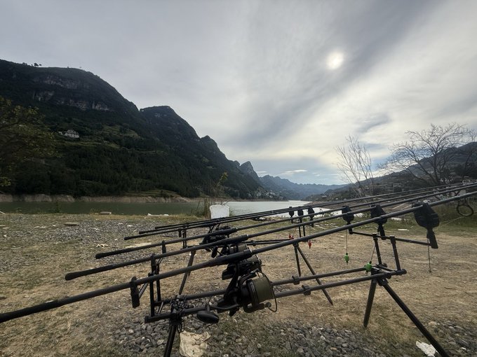A landscape view of a fishing setup with multiple rods on stands by a lake, with a small boat on the water and mountains in the background under a hazy sky.