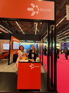 The image shows a convention center with two women standing next to an orange booth displaying the company's logo, which reads "QUANT". The booth appears to be part of a larger event or exhibition taking place at the convention center. There are other people in the background, indicating that it is a busy and lively gathering. The post about the event starts on Day 1 and continues until Day 2, suggesting that there might be more activities or discussions happening during this time frame.
