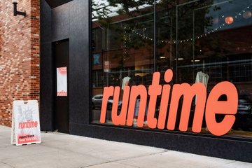 The storefront of a 'runtime' restaurant with bright orange signage on the windows and a sandwich board sign out front.