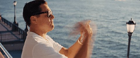 The image shows a man wearing glasses standing next to a body of water near the ocean. He appears to be at a pier or dock area by the water, with a cityscape visible in the background. The atmosphere seems relaxed as he enjoys his time by the sea.
