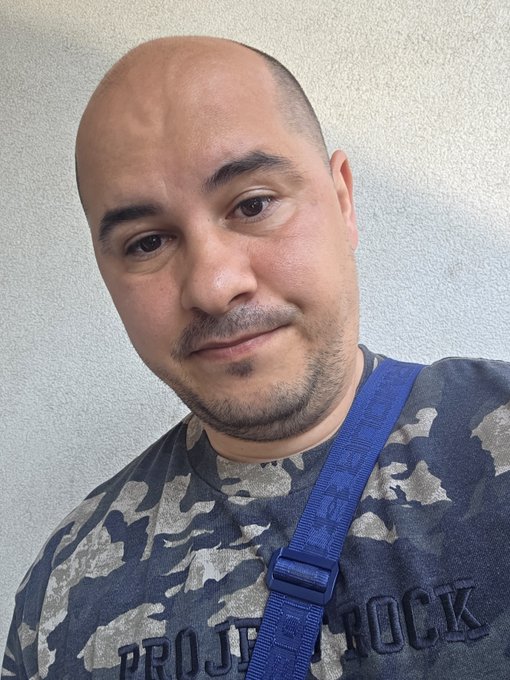 A bald man with a goatee takes a selfie wearing a gray camouflage shirt that reads "PROJECT [partially obscured]" and a blue crossbody strap, looking unenthusiastic—matching the post's relatable sentiment about being unhappy that vacation is over and dreading an early gym session.
