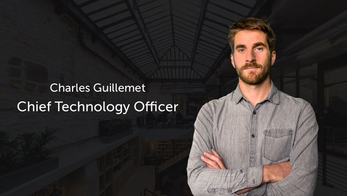 A professional headshot of Charles Guillemet, Chief Technology Officer, a man with a beard wearing a gray button-up shirt, standing with arms crossed in a modern office setting.