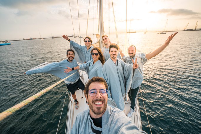 A group of friends in matching light blue shirts pose enthusiastically for a selfie on a sailboat during a golden sunset on the water.