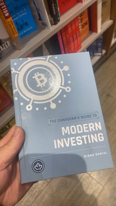 A hand holding a light blue book titled 'The Canadian's Guide to Modern Investing' by Kiana Danial, featuring a Bitcoin logo, in front of a bookstore shelf.