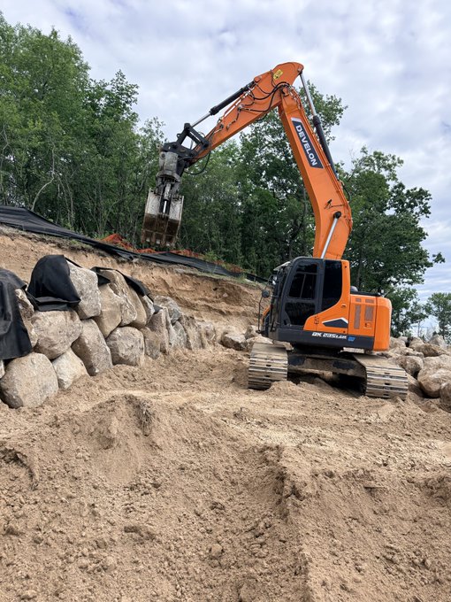 The image shows a large orange construction vehicle with an excavator arm lifting up rocks from the ground. The scene takes place outdoors, possibly at a construction site or a rocky hillside where the vehicle is working. There are several rocks scattered around the area, indicating that the vehicle has been actively digging and moving them.