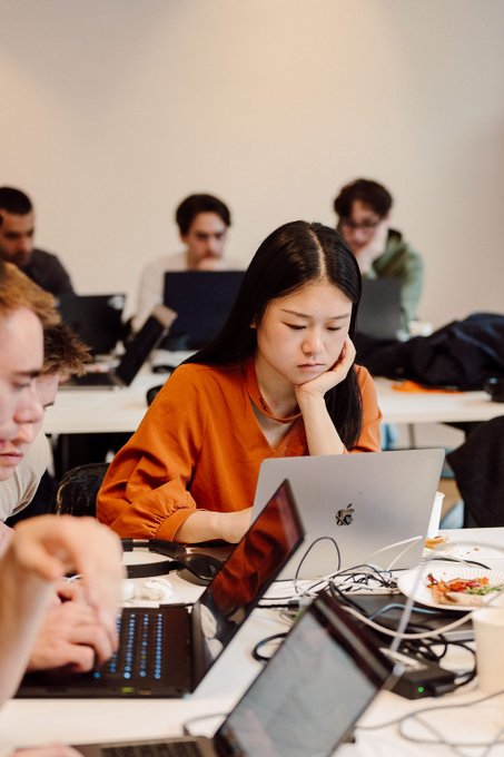A young woman with long dark hair in an orange shirt rests her chin on her hand while looking at a silver laptop in a room filled with other people working on computers.