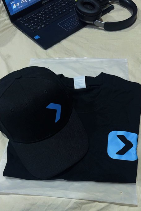 A black cap and t-shirt featuring the blue Bitget logo (a stylized ">" arrow) are displayed on a desk next to a laptop and headphones, representing a promotional merchandise package received from Bitget, thanking @Bigkhaddyy for the delivery.