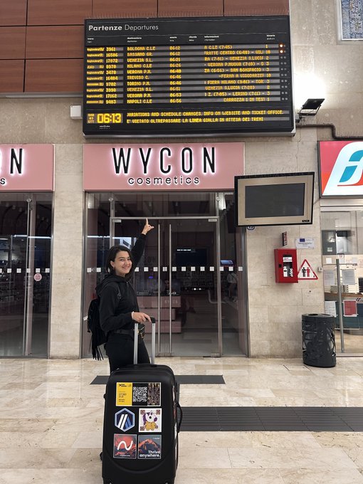 A person in a black jacket stands at a train station below a departures board showing the time 06:13, holding the handle of a sticker-covered suitcase in front of a 'WYCON cosmetics' store.