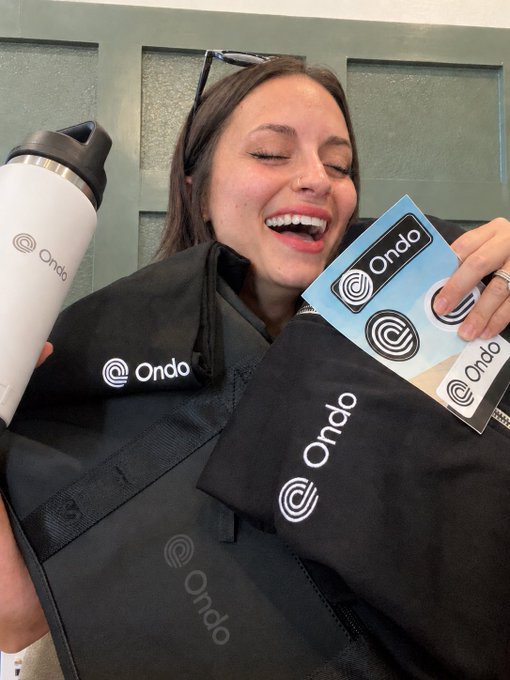 A smiling woman holding Ondo-branded merchandise including a water bottle, stickers, and a jacket/bag.