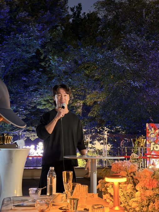 A man in black clothing speaking into a microphone at an outdoor evening event, with trees illuminated in blue/purple light behind him and a table with drinks and orange flowers in the foreground.