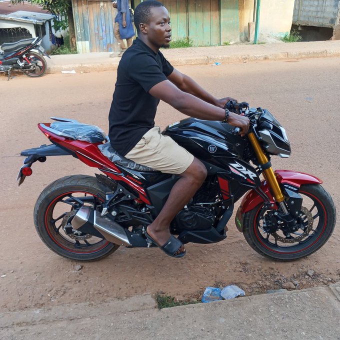 The image shows a man sitting on a motorcycle, wearing black clothing and tan shorts. The motorcycle has red accents and is parked next to a curb. There are two other motorcycles visible in the background, one of which appears to be a GM Ct.