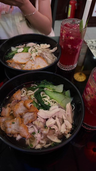Two large black bowls filled with noodle soup topped with pork, chicken, and greens, served alongside tall glasses of red iced drinks.